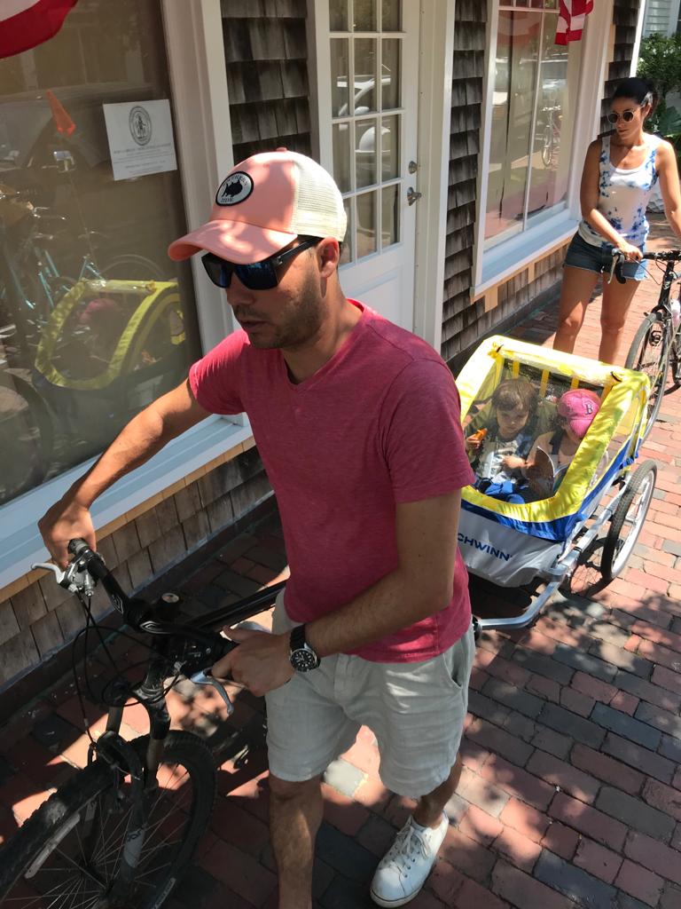 A man wearing a hat and a red t-shirt is pushing a bike with a kids trailer. behind a woman is pushing a walking a bike.