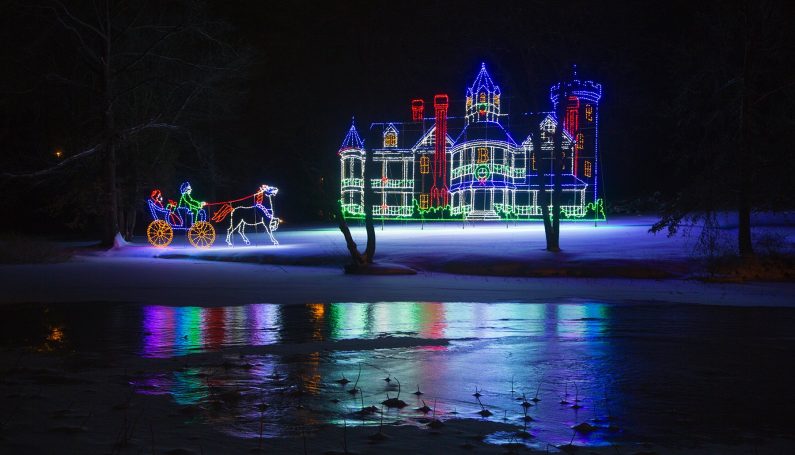 An outline of a Victorian home in Christmas lights with an outline of a horse and carriage in Christmas lights to the left. Ice and snow in front.