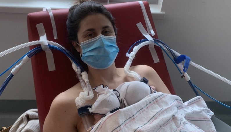 A mom wearing a face mask while holding premature twins in a chair connected to multiple medical devices (Prematurity Awareness Month)