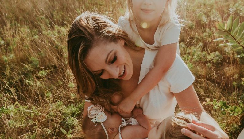 Mother kneels and nurses baby with two other young daughters surrounding her ("let them" theory for parents)
