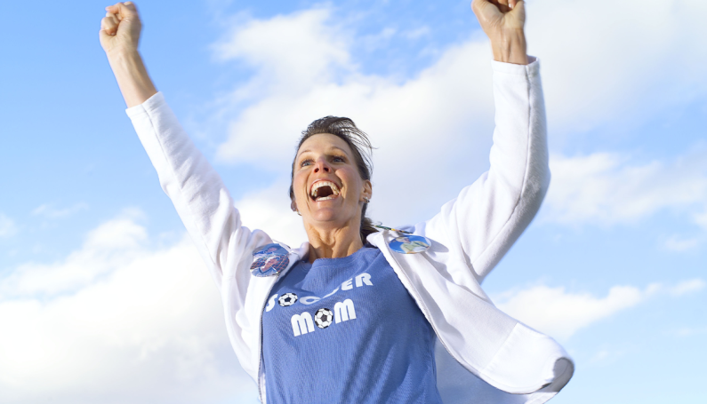 woman with blue "soccer mom" shirt raising her hands in the air, cheering