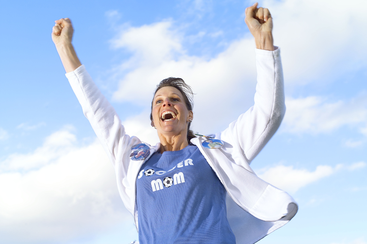 woman with blue "soccer mom" shirt raising her hands in the air, cheering