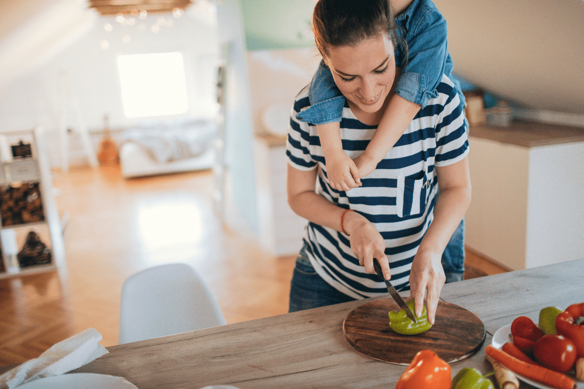 mom chopping peppers with a child hanging on her back, illustrating the idea of a monthly family meal plan