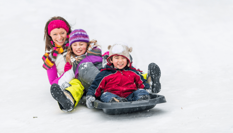 mother and two children sitting together on a gray sled on a snowy hill
