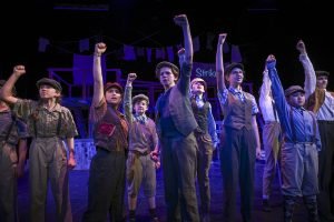 Boys on stage in a production of Newsies. They are wearing newsboy outfits and they each have one arm raised. 