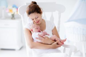 Young mother holding her newborn child. Mom nursing baby. Woman and new born boy relax in a white bedroom with rocking chair and blue crib. Nursery interior. Mother breast feeding baby. Family at home