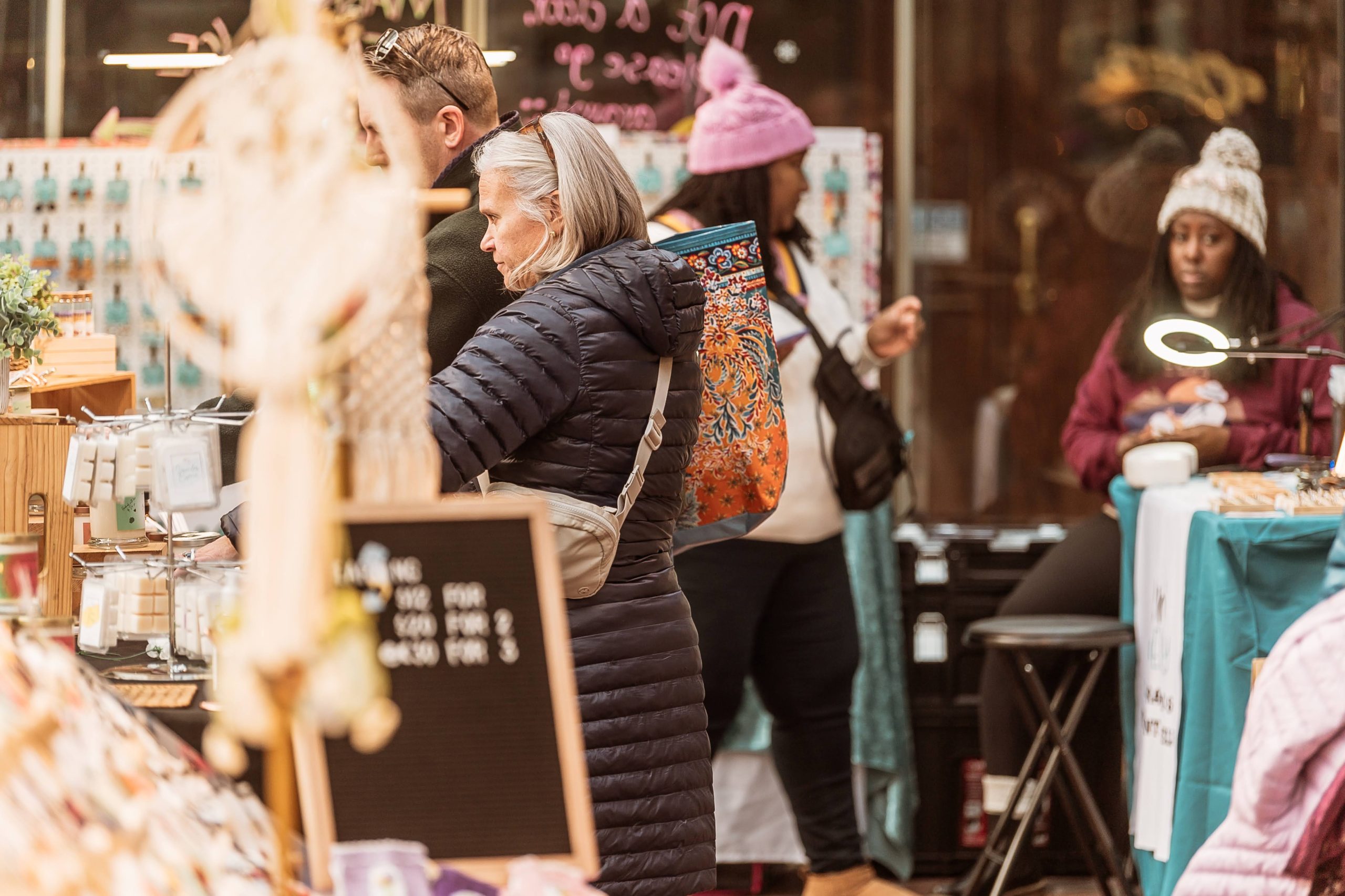 Woman with shoulder length gray hair and a long black winter coat shopping at an indoor holiday market. Two women in winter hats in the background. A dreamcatcher hanging on the left side.