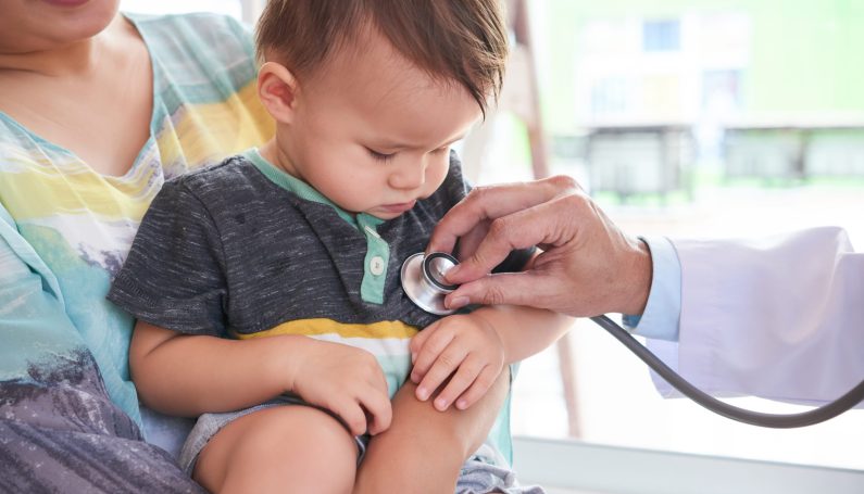 Cute toddler sitting on his mother's knees while doctor listening to his heart beat