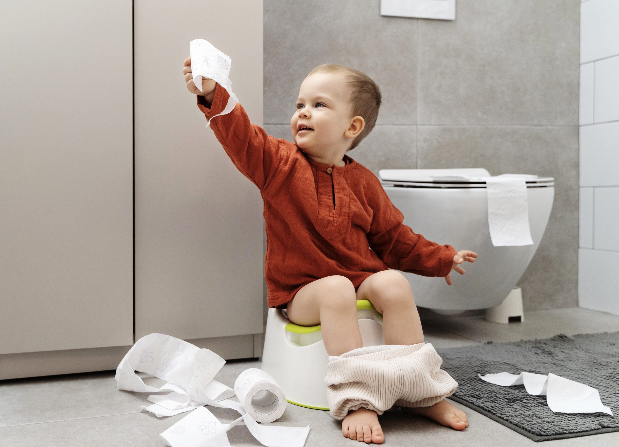 toddler in red shirt holding toilet paper and sitting on small potty learning to potty train