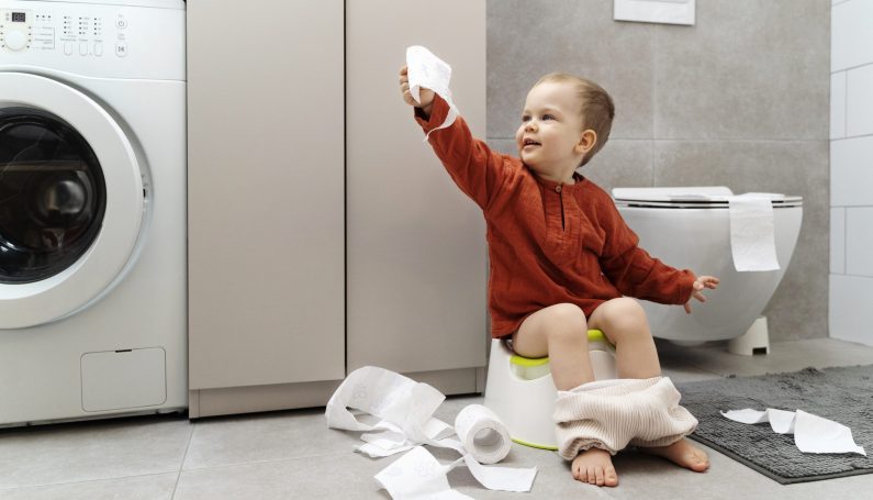 toddler in red shirt holding toilet paper and sitting on small potty learning to potty train