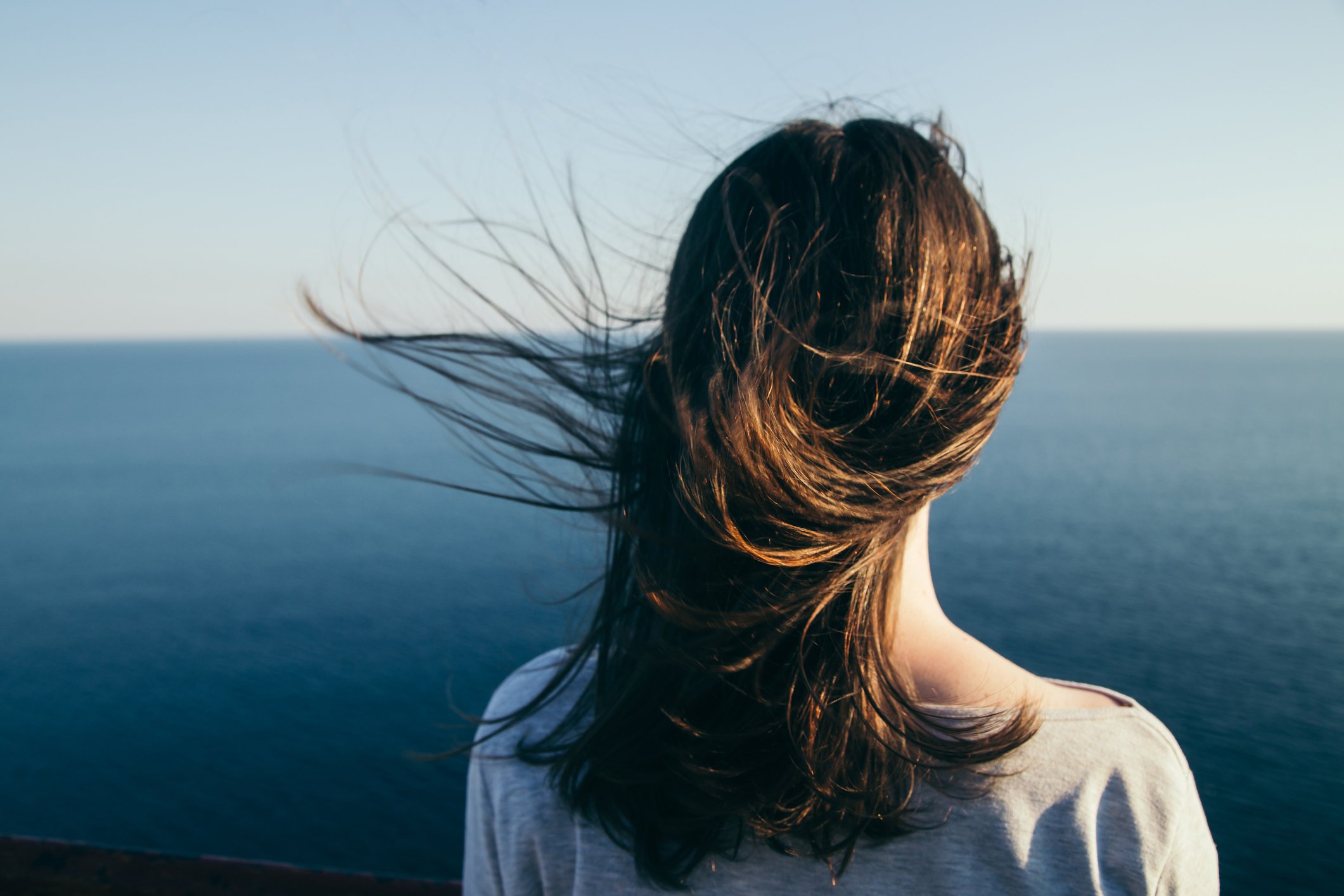 Woman with dark hair stands on a top cliff over blue sea view while wind. (stopped breastfeeding to start mammograms for breast cancer)