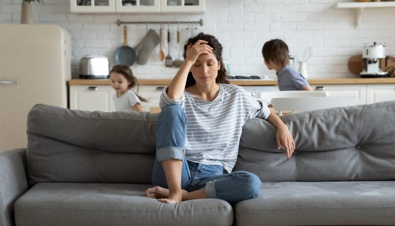 Tired mother suffers from headache, closed eyes, touching forehead, sitting on couch while her children run around her, first trimester pregnancy sickness