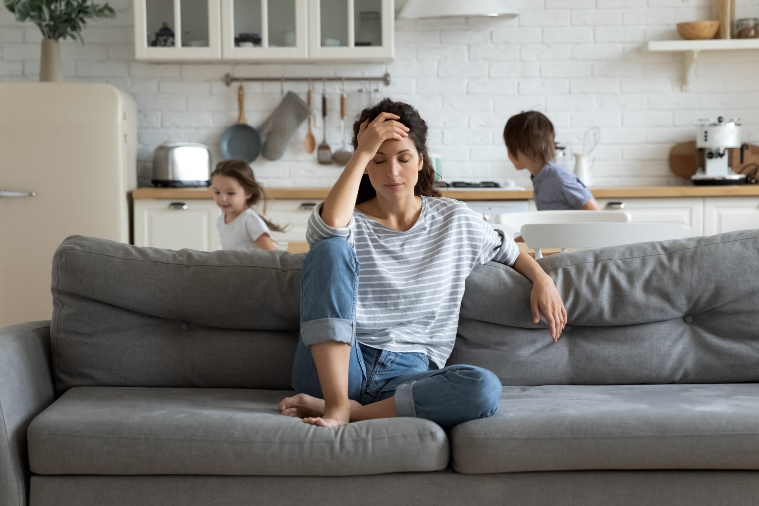 Tired mother suffers from headache, closed eyes, touching forehead, sitting on couch while her children run around her, first trimester pregnancy sickness