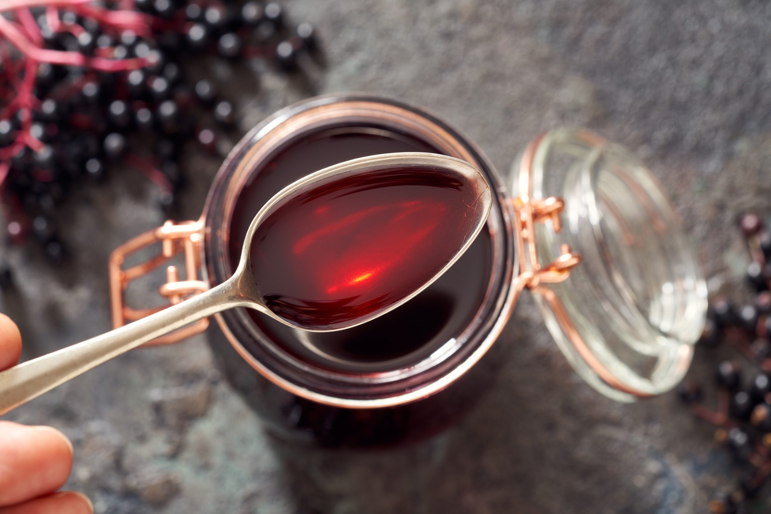 Spoon with homemade elderberry syrup, with fresh elderberries in the background (cold and flu home remedies)