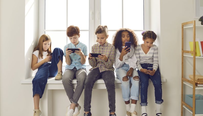 group of kids sit in a row on a windowsill staring at smartphones (Wait Until 8th, delaying technology for kids and smartphones)