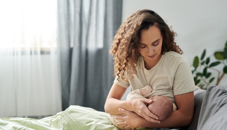 Young mother in pajamas sitting under blanket on bed breastfeeding her baby
