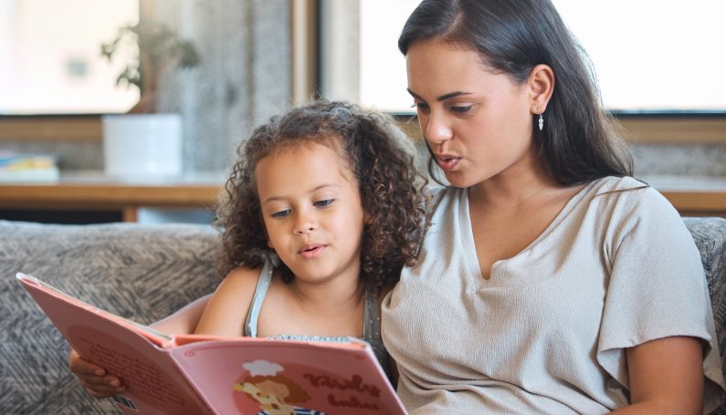 Loving hispanic mother and her little daughter sitting at home and reading a storybook together. raising bilingual children