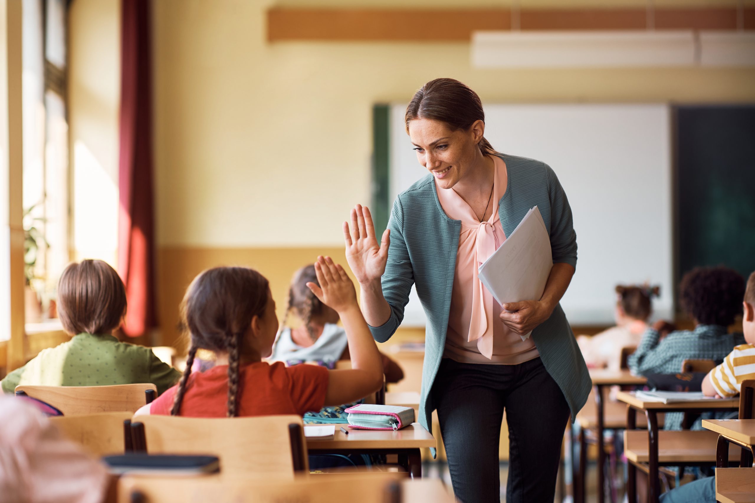 Happy elementary school teacher giving high-five to her student during class in the classroom. (teacher and students, school supplies, tissues)