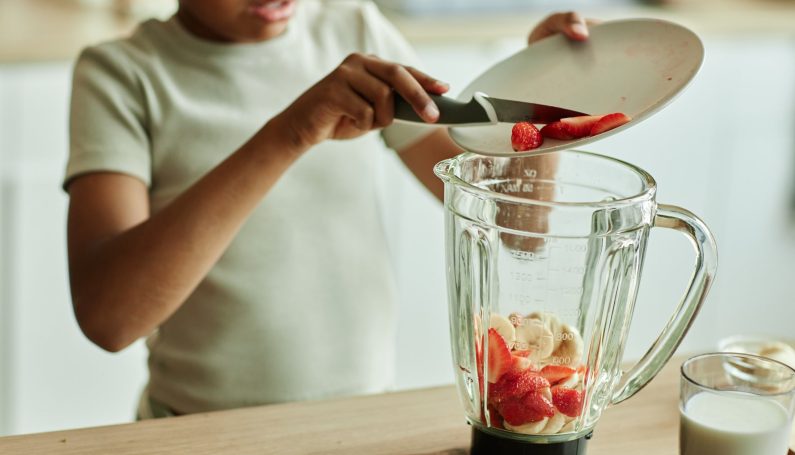 little girl carefully adding strawberries to blender making fruit smoothie with banana (snacks kids can make on their own)