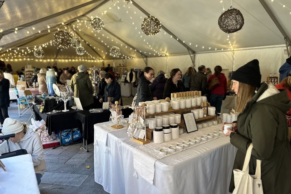 People shopping at an indoor holiday market located inside a large, white, canvas tent with hanging string lights and round balls covered in lights hanging from the ceiling.