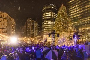 A large crowd of people outside at night with a large lit-up Christmas tree in the background, along with tall buildings with lights on inside.