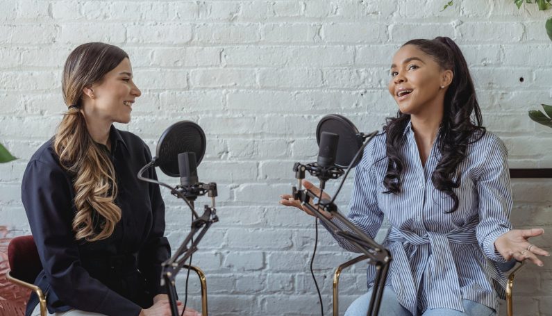 two women speaking into microphones, recording a podcast episode (podcasts for busy working moms)