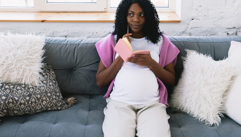 pregnant woman sitting on couch with pen and notebook in hand, thinking about what to choose for her child's last name