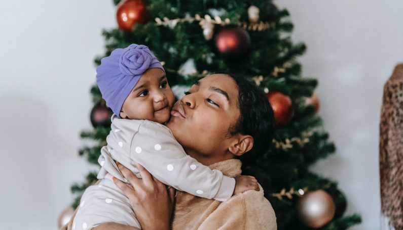 Black woman holding and kissing a baby in front of a Christmas tree (holidays with newborn baby)