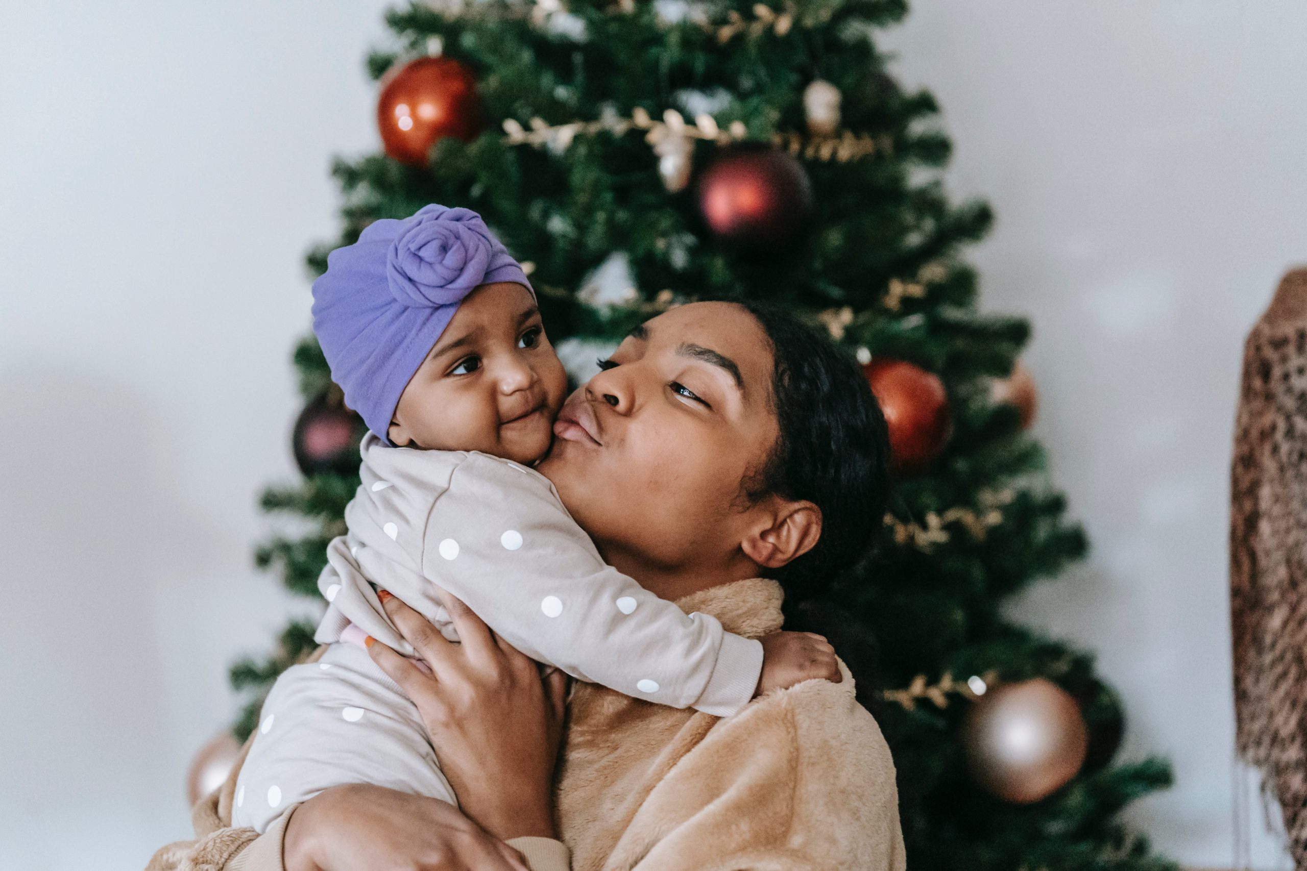 Black woman holding and kissing a baby in front of a Christmas tree (holidays with newborn baby)