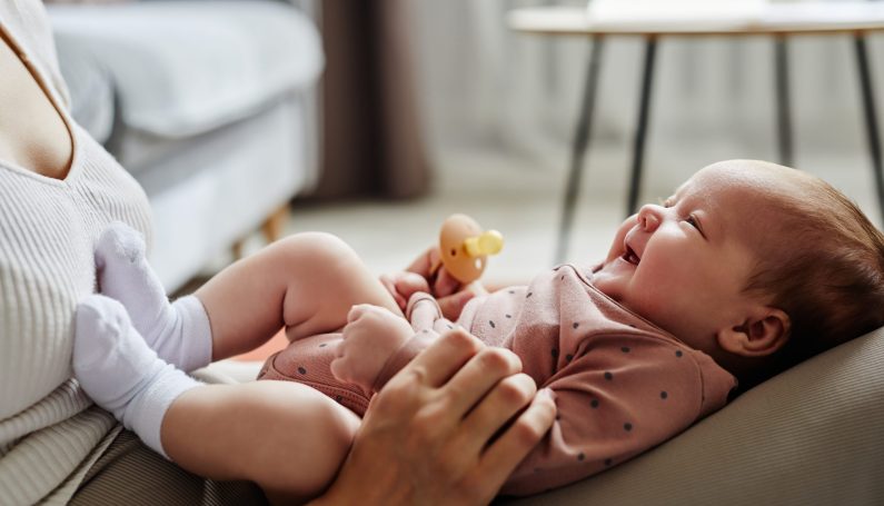 Side view of mother playing with laughing baby girl lying on her knees spending time at home (New England postpartum doula support)